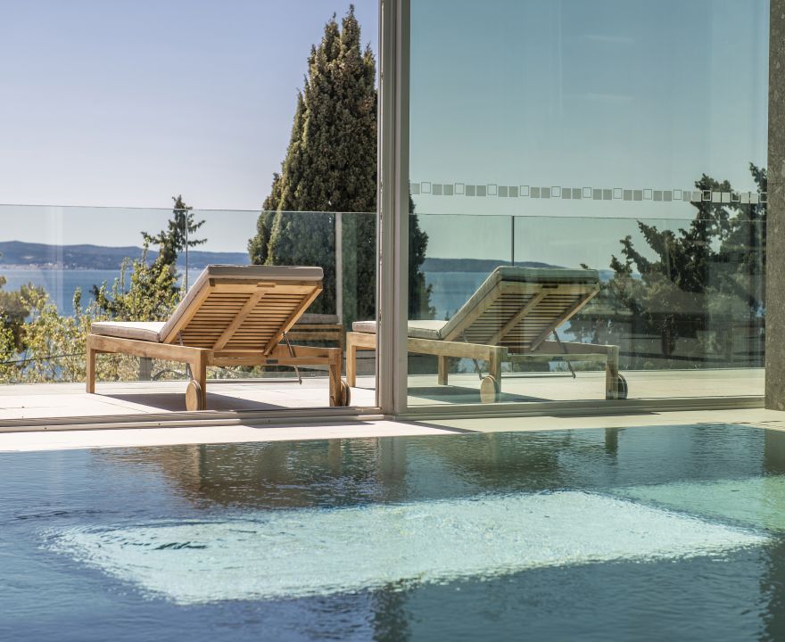 Man sieht einen Pool mit türkisem Wasser. Im Hintergrund sind Holzliegen zum Entspannen. 